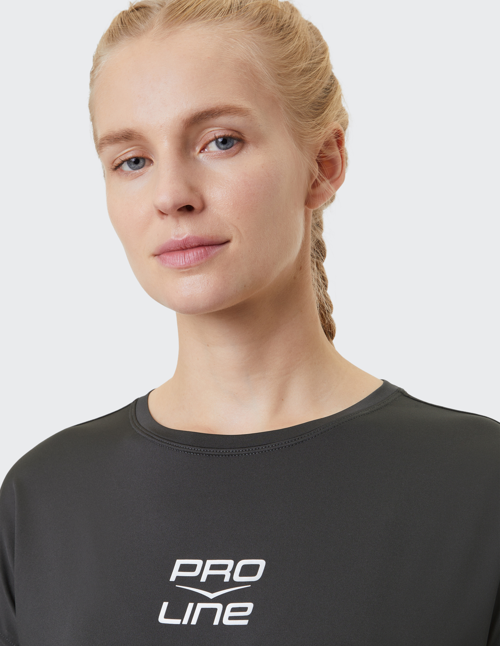 Frau mit geflochtenem Haar in schwarzem Shirt mit weißem "PRO LINE"-Logo auf hellgrauem Hintergrund, blickt freundlich in die Kamera.