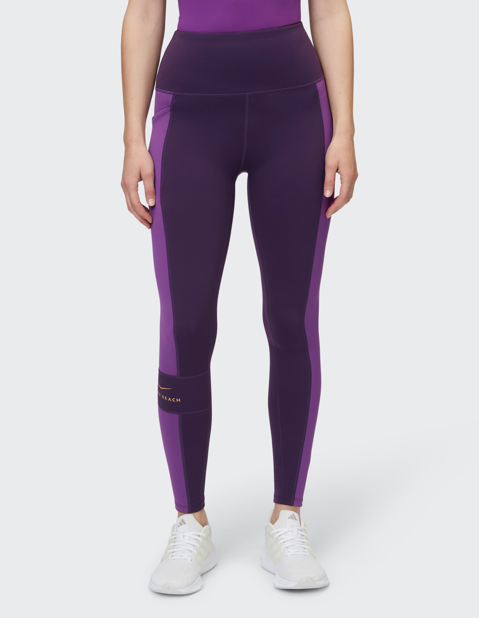 Frau trägt eine zweifarbige, violett-lila Leggings mit hohem Bund und weißen Turnschuhen, vor neutralem Hintergrund.