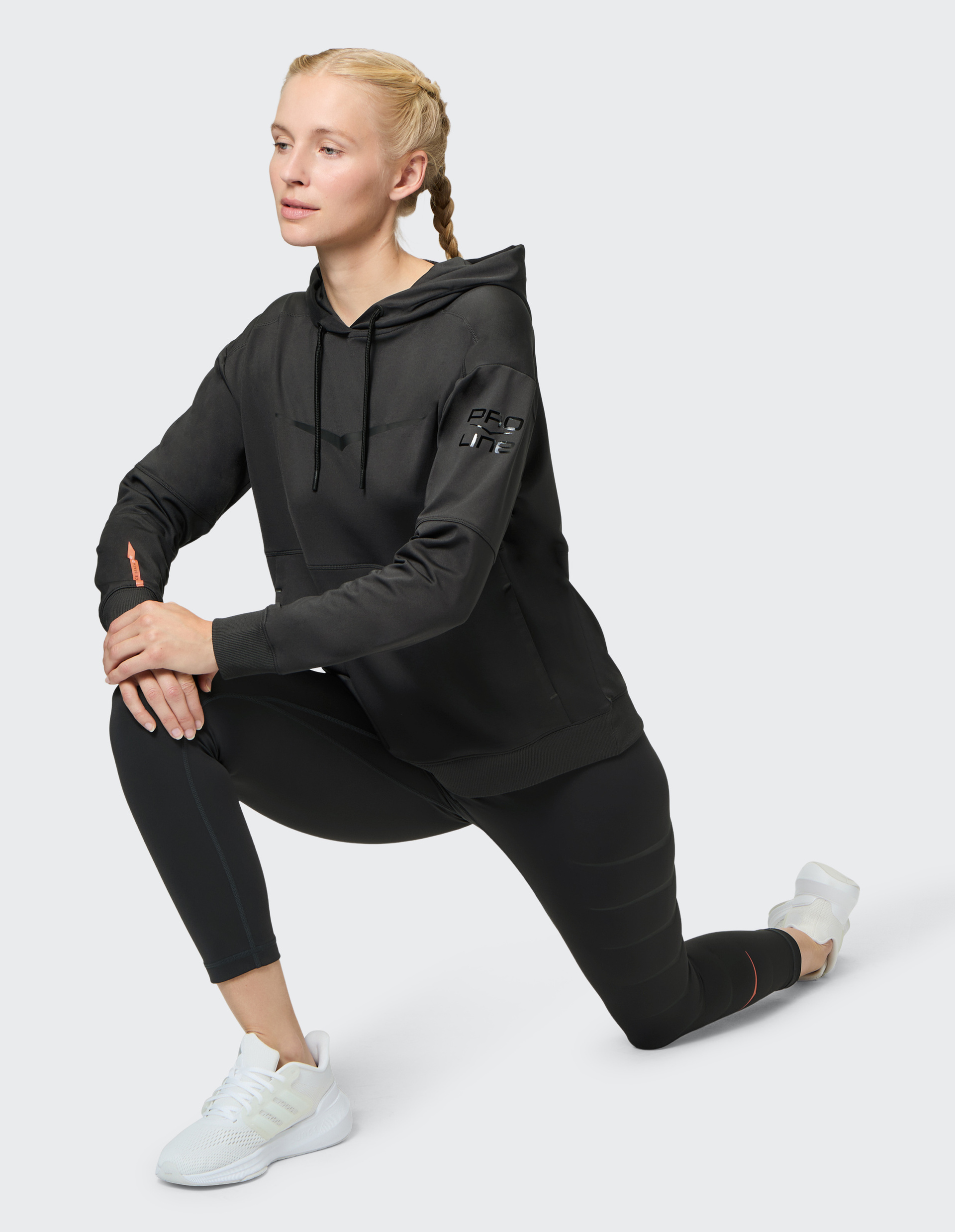 Frau mit blonden Zöpfen macht in schwarzem Sportanzug mit Kapuze und weißen Turnschuhen einen Ausfallschritt, auf hellem Hintergrund.