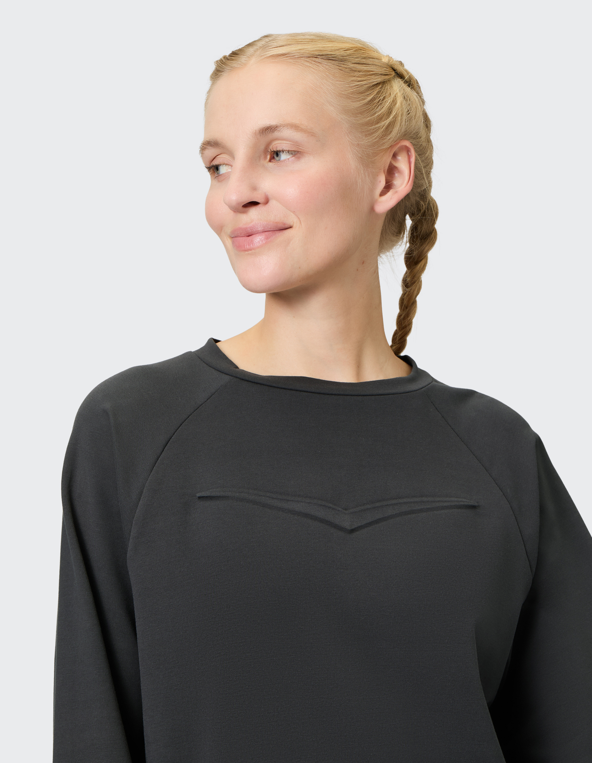 Frau mit blondem Zopf trägt dunkelgraues Oberteil, schaut lächelnd zur Seite, vor hellem Hintergrund.
