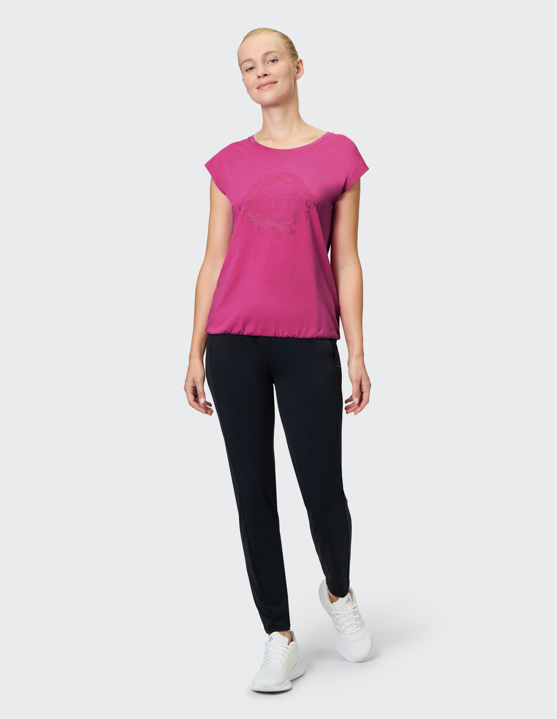 Frau in pinkem T-Shirt mit "Venice Beach"-Aufdruck, schwarzen Leggings und weißen Sneakers auf hellem Hintergrund lächelt leicht.