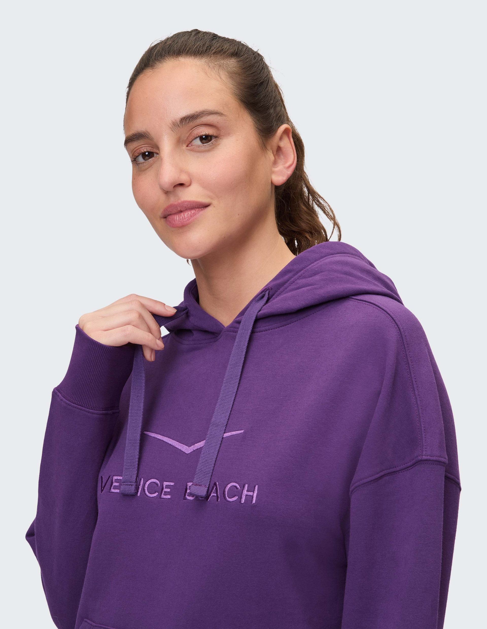 Frau lächelt leicht und trägt einen lila Hoodie mit "Venice Beach"-Aufdruck, ihr Haar ist zu einem Zopf zurückgebunden.