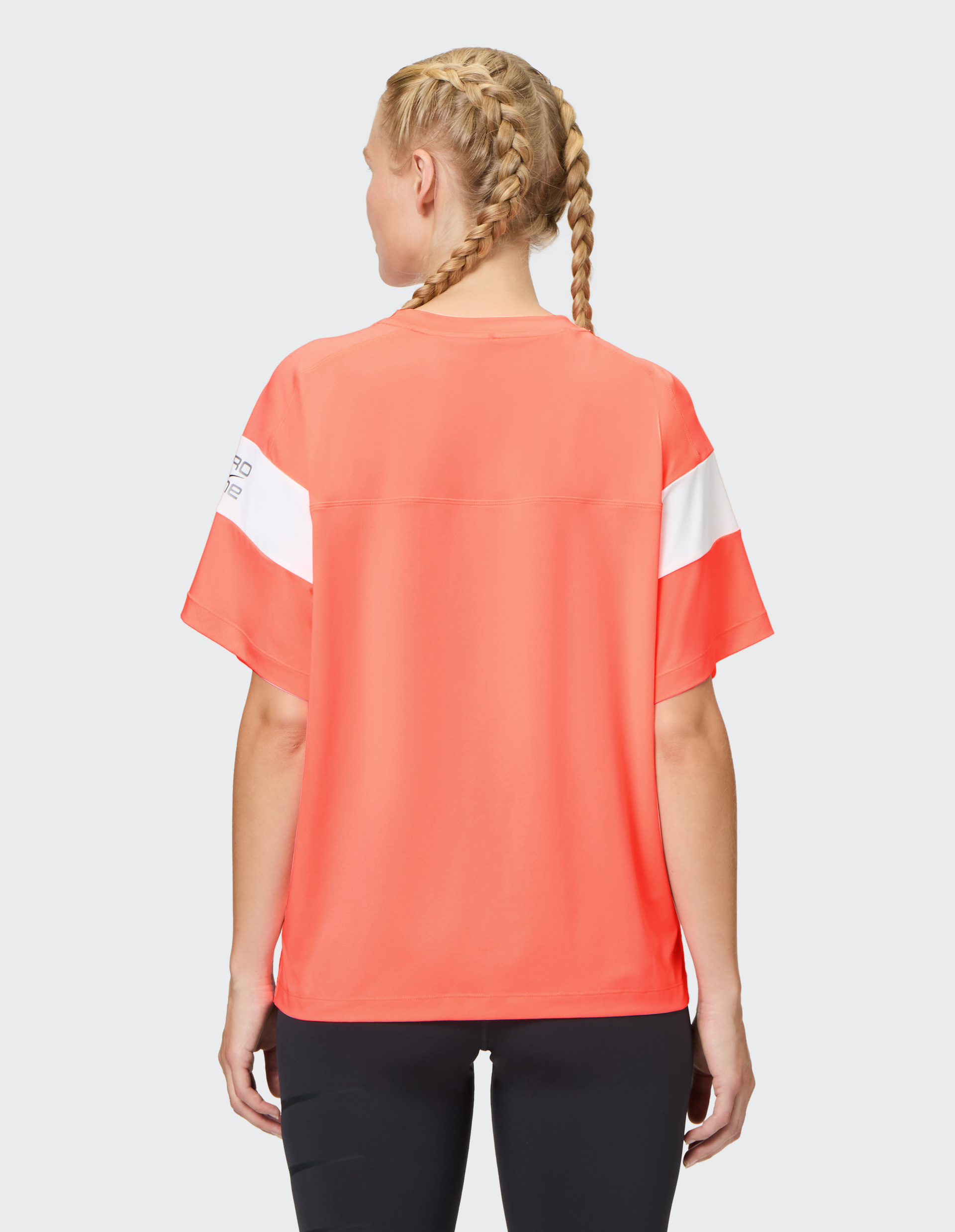 Frau mit geflochtenem Haar trägt ein korallenrotes T-Shirt mit weißem Streifen auf dem Ärmel, steht rückwärts, schwarze Leggings.