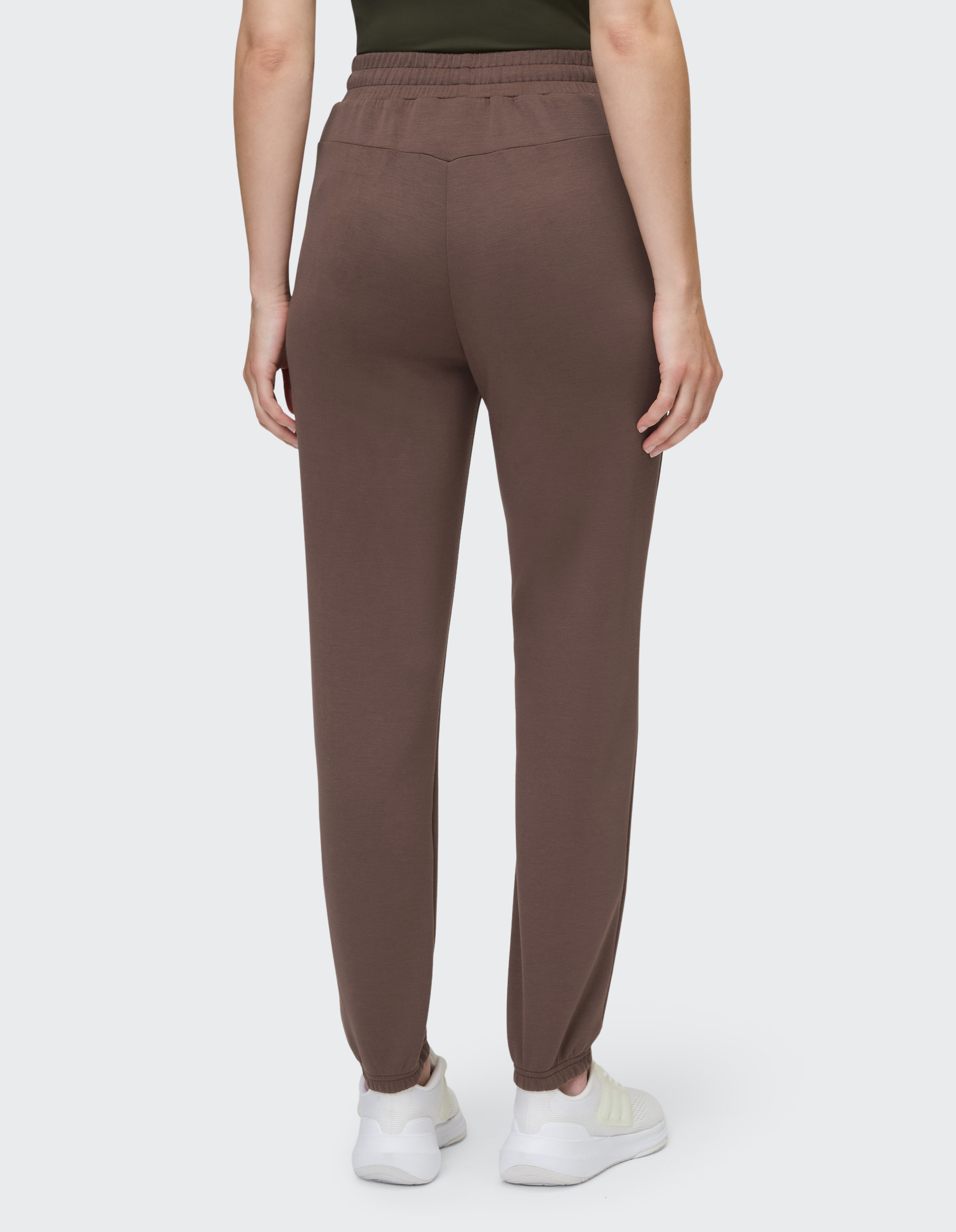 Rückansicht einer Person in brauner Jogginghose mit Gummibund und weißen Sneakern vor grauem Hintergrund.