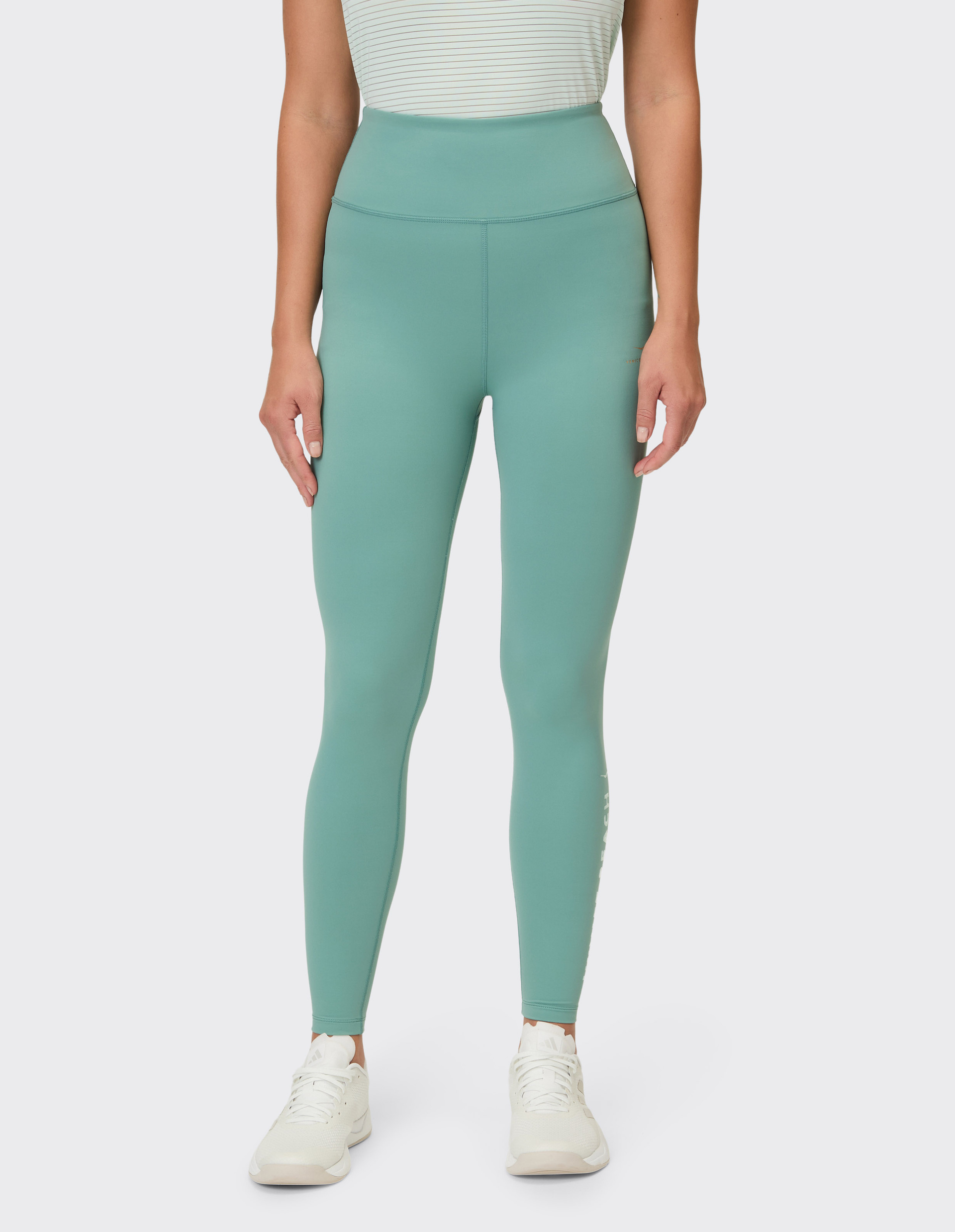 Eine Frau trägt schlichte, türkisfarbene Leggings mit dezentem Logo am linken Bein und weiße Turnschuhe.