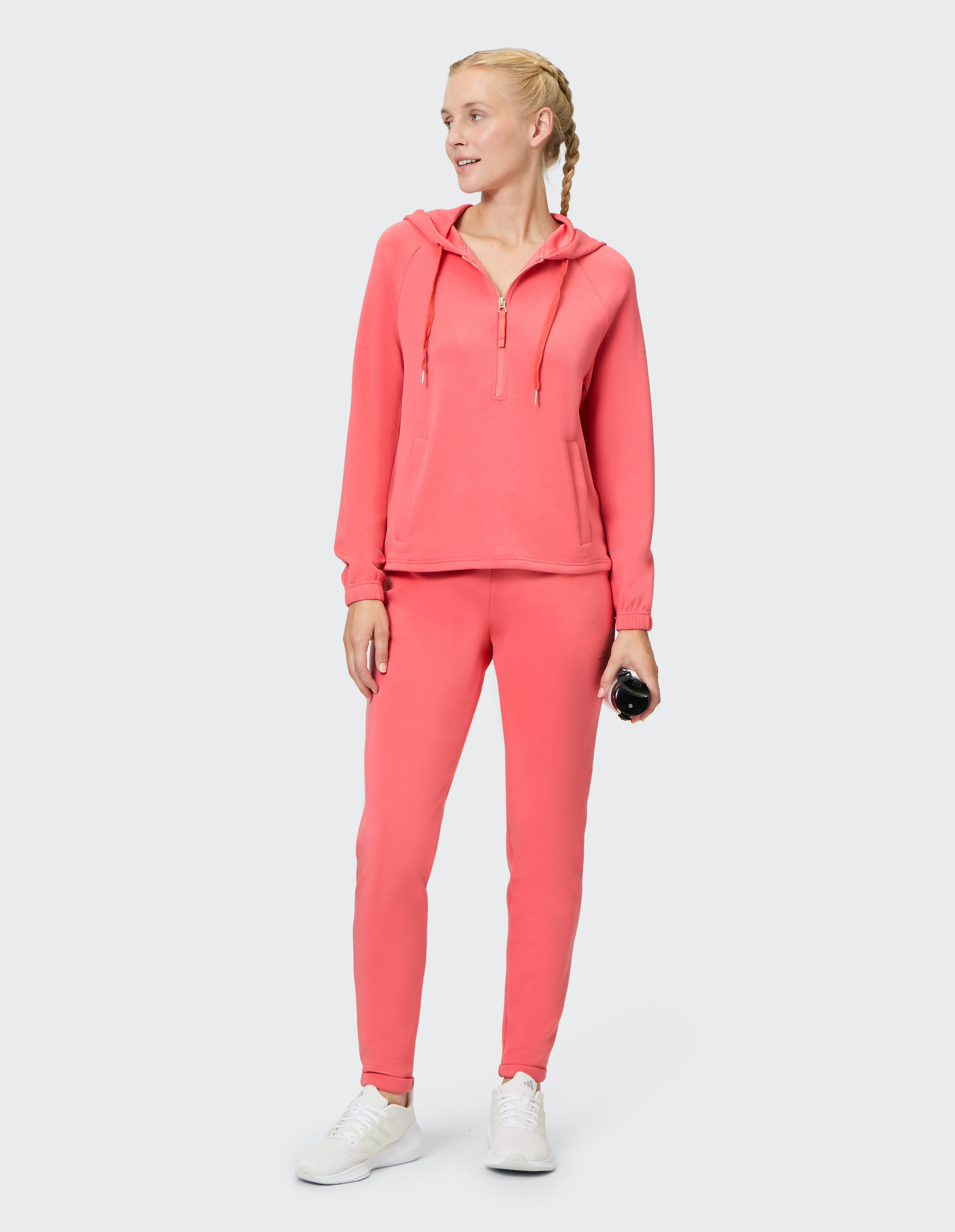 Frau in korallfarbenem Jogginganzug und weißen Sneakers hält eine schwarze Trinkflasche, lächelnd nach rechts schauend.