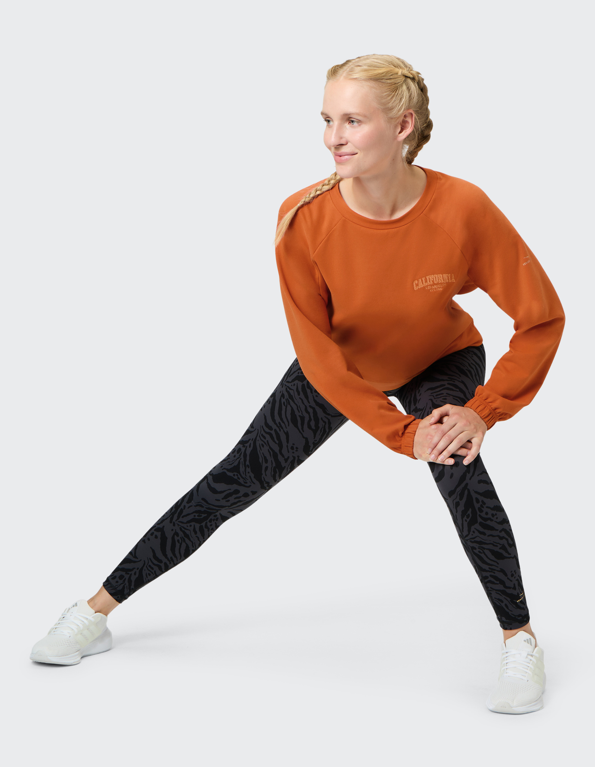 Eine Frau in orangefarbenem Pullover und gemusterten Leggings dehnt sich, lächelnd in sportlicher Pose, auf grauem Hintergrund.