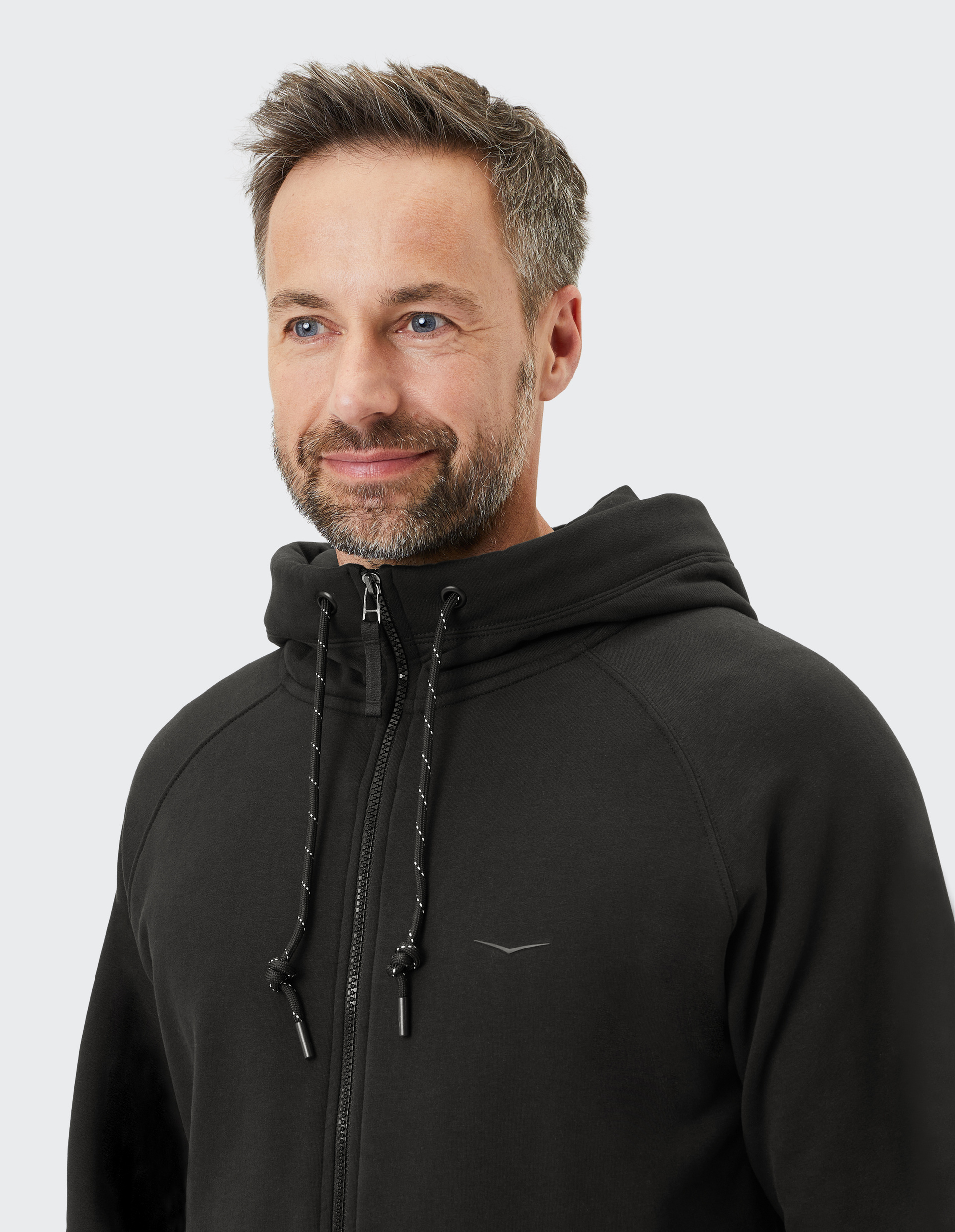 Ein lächelnder Mann mit Bart trägt einen schwarzen Hoodie mit Kordelzug und kleinen Logo auf der Brust vor grauem Hintergrund.