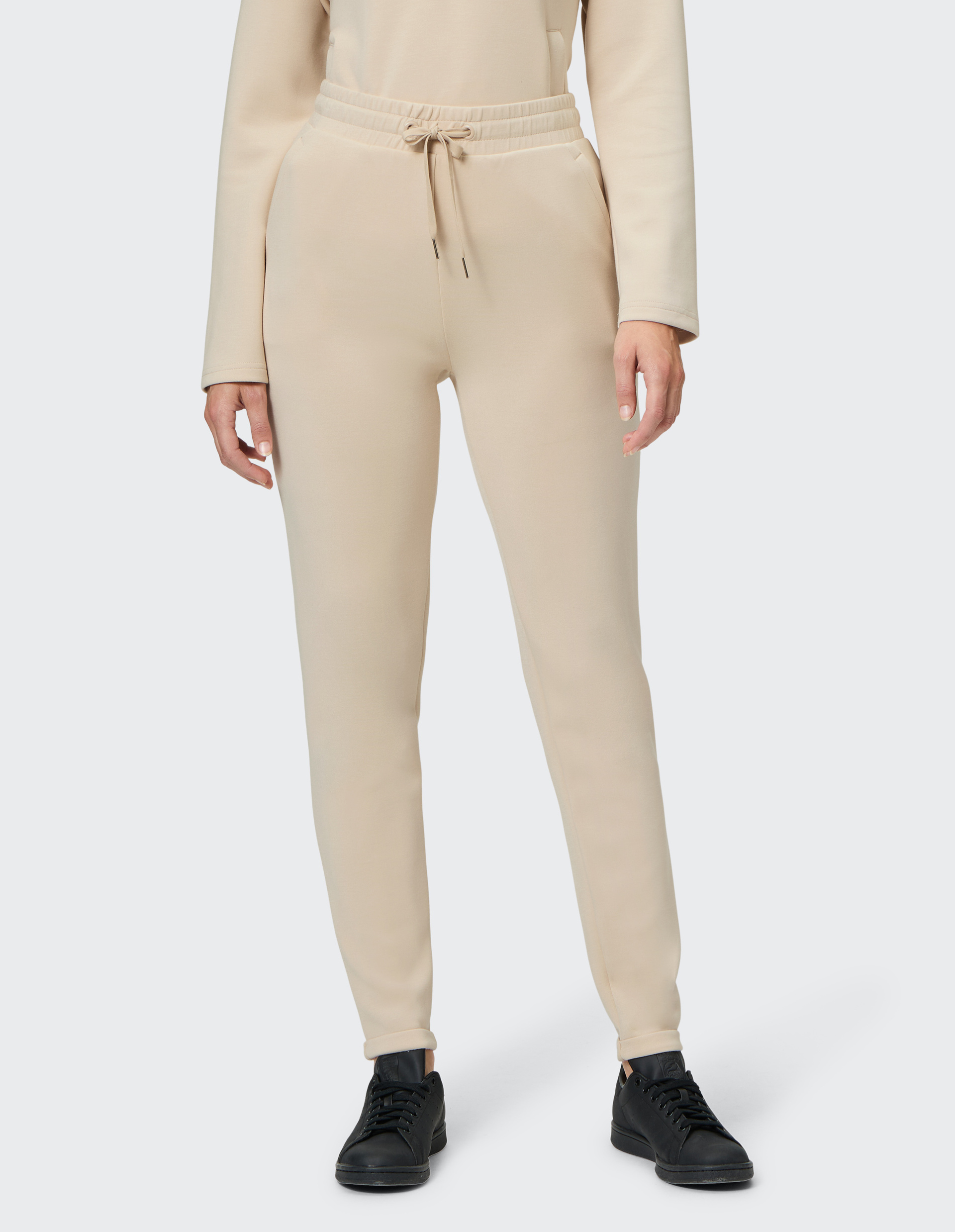 Eine Person trägt eine beige Jogginghose mit Kordelzug und schwarzen Sneakers, das Oberteil ist gleichfarbig und langärmelig.