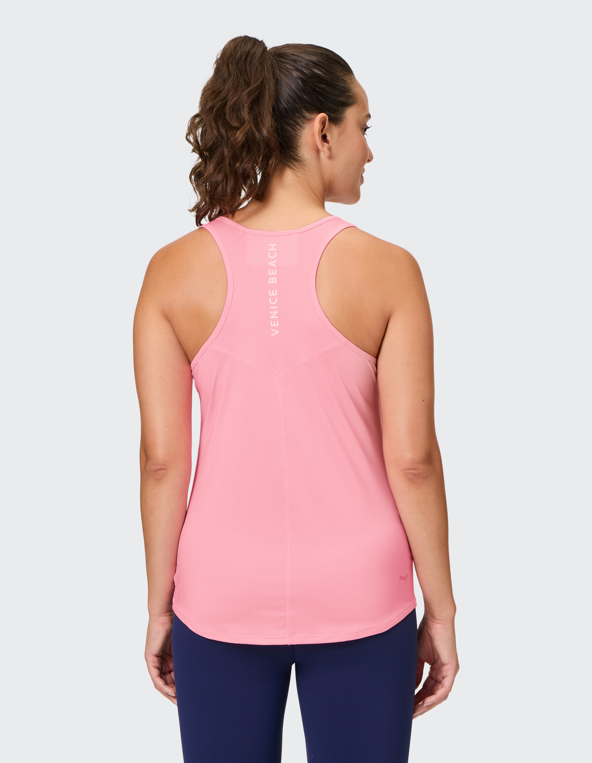 Frau von hinten in pinkem Sporttop mit Aufschrift "Venice Beach", dunkelblauer Leggings und lockigem Pferdeschwanz, auf grauem Hintergrund.