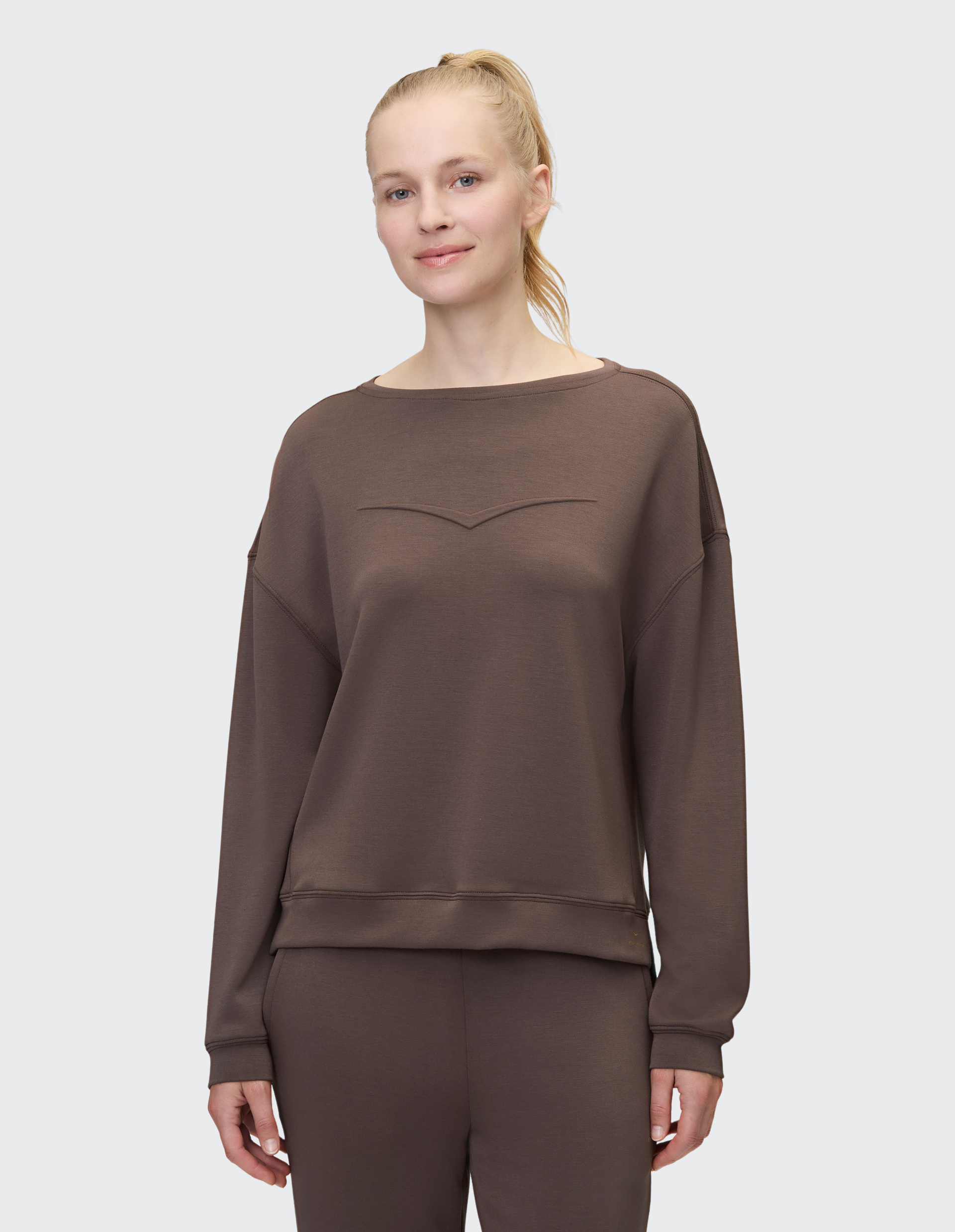 Frau mit lächelndem Gesicht in braunem Sweatshirt und passender Hose, blondes Haar in einem Pferdeschwanz, vor grauem Hintergrund.