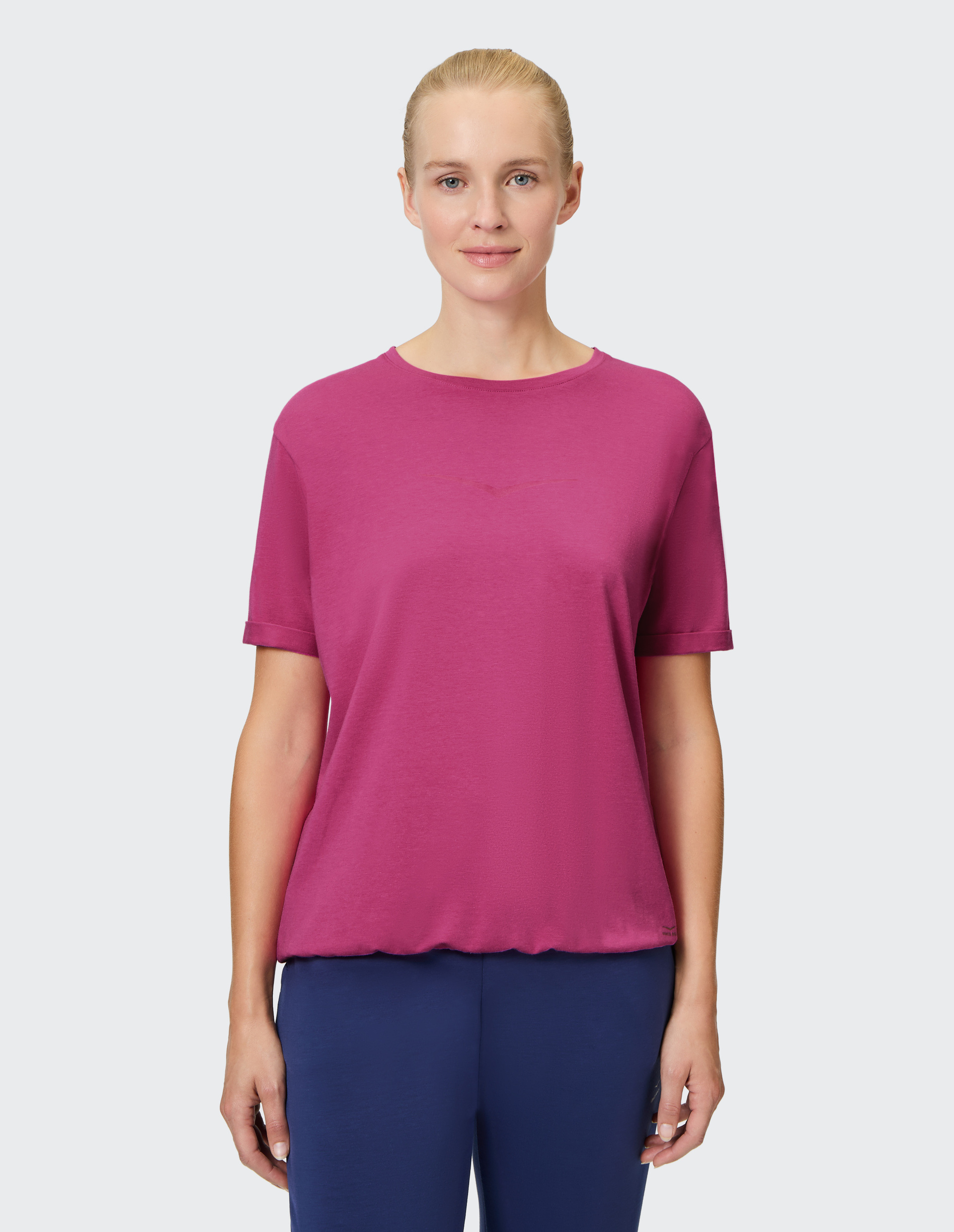 Person in pinkem T-Shirt und blauer Hose steht vor grauem Hintergrund und lächelt leicht in die Kamera.