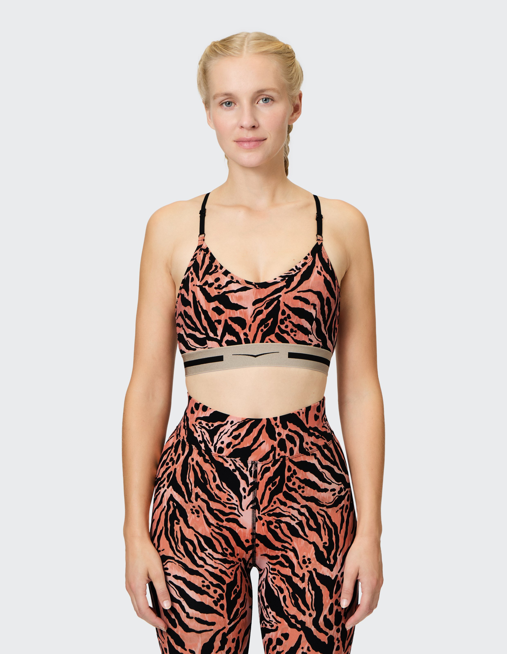 Eine Frau mit geflochtenem Haar trägt ein sportliches, orange-schwarzes, zebragemustertes Fitness-Outfit vor einem hellgrauen Hintergrund.