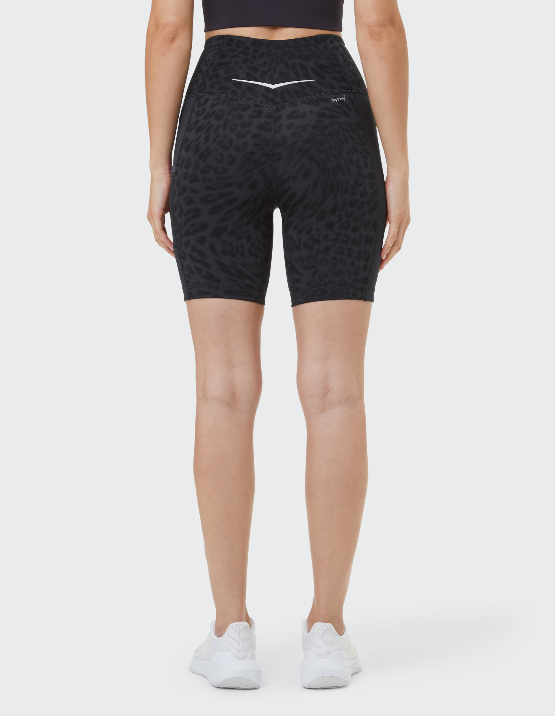 Frau in schwarzen, gemusterten Radlershorts und weißen Sneakers, von hinten fotografiert, in neutralem Raum.