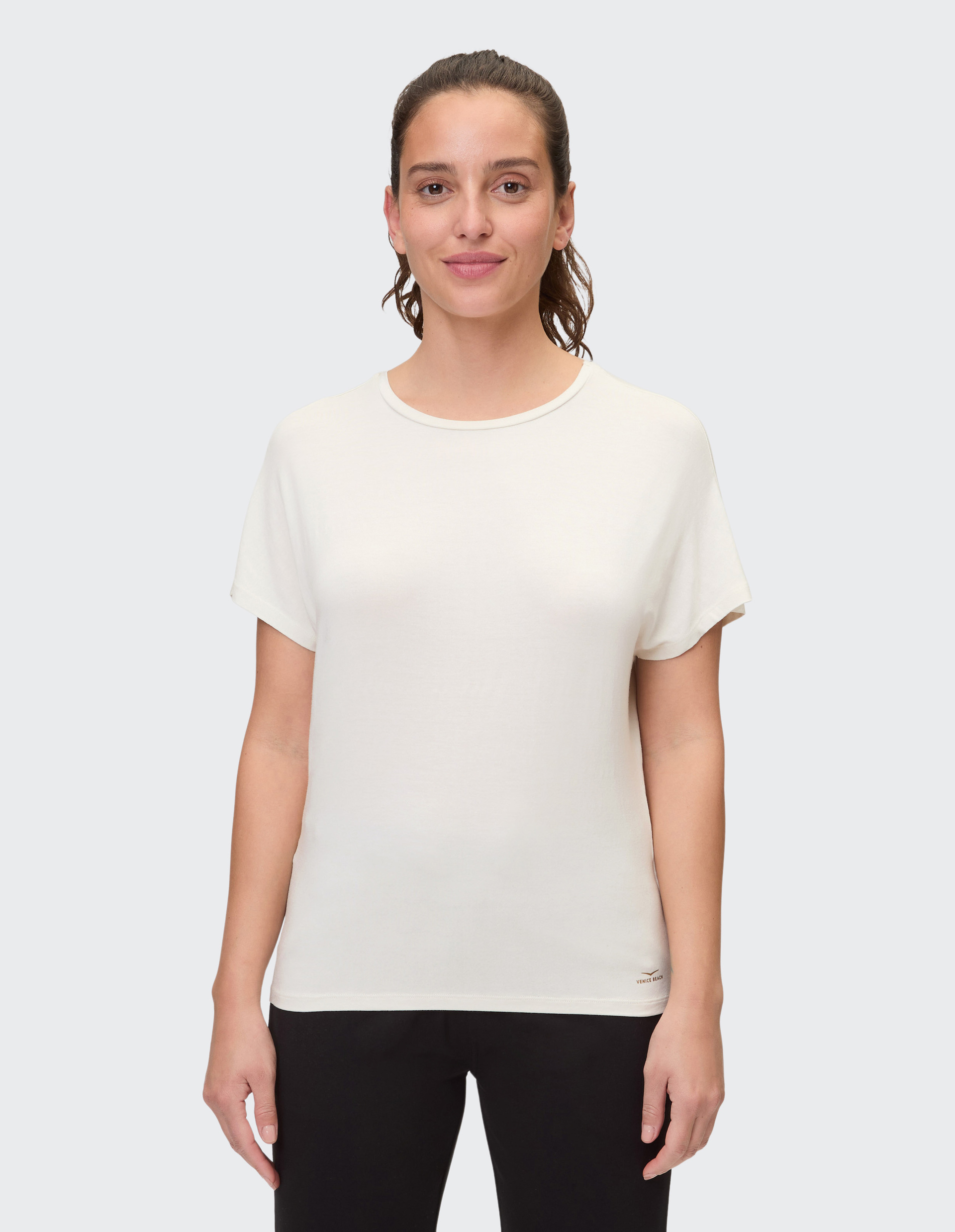 Frau mit halblangen Haaren trägt ein weißes T-Shirt und schwarze Hose, lächelt leicht vor neutralem Hintergrund.
