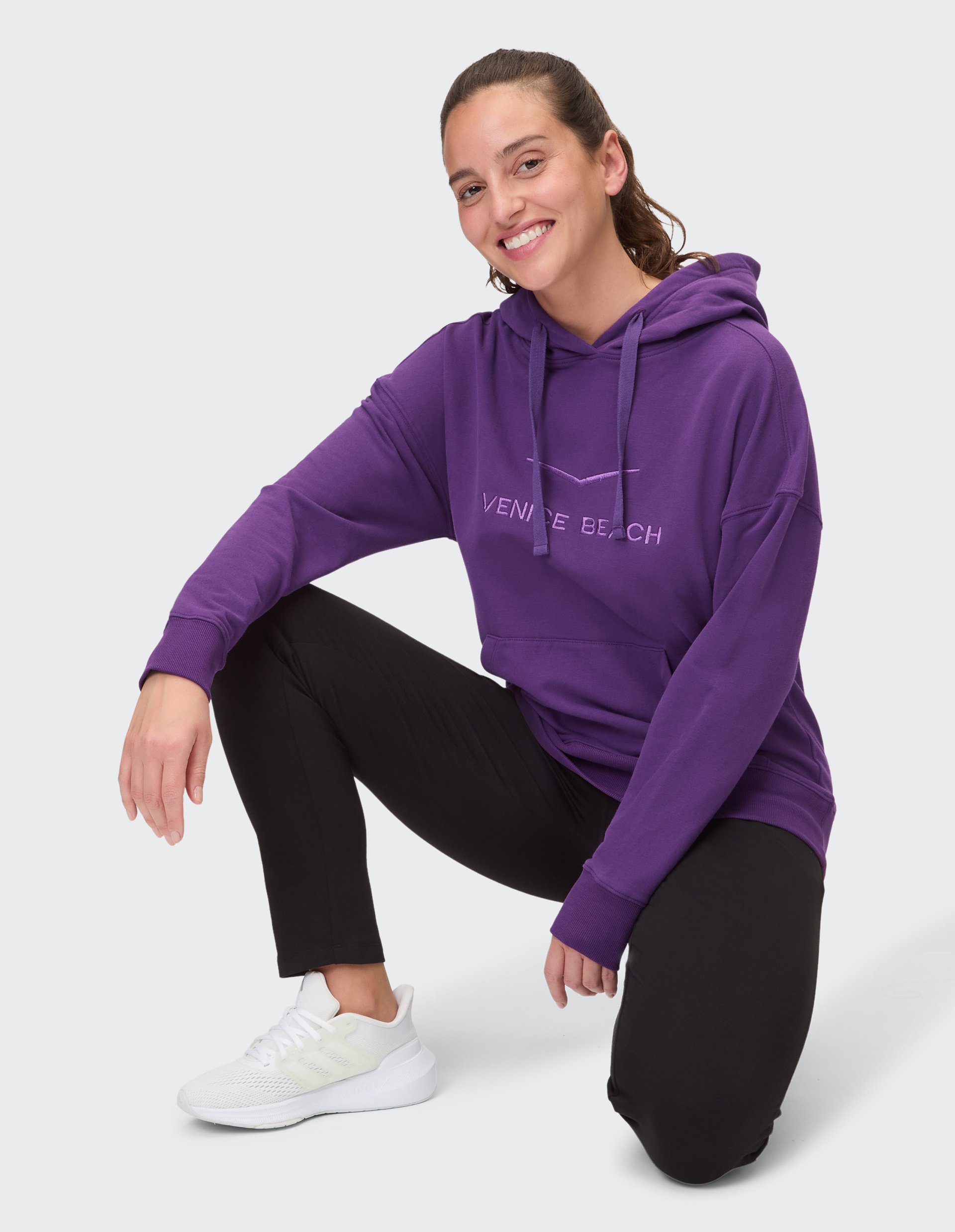 Person in lila Hoodie mit "Venice Beach"-Schriftzug, schwarzen Leggings und weißen Turnschuhen vor neutralem Hintergrund.