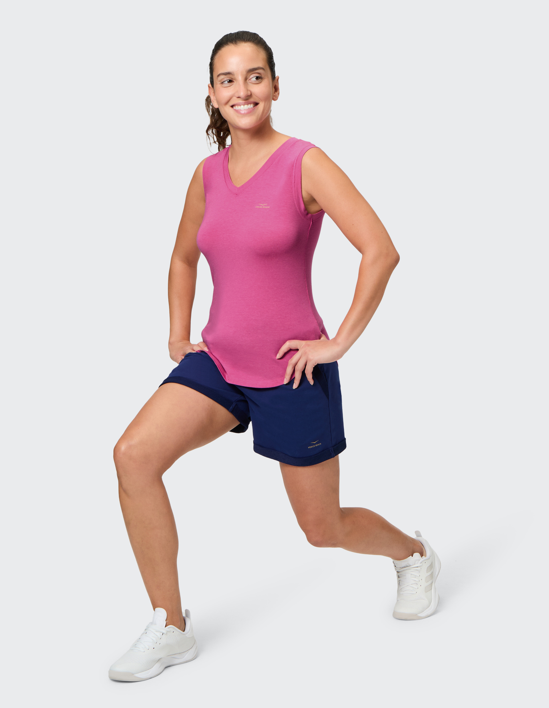 Frau im pinken Sporttop und blauen Shorts macht einen Ausfallschritt, lächelnd, mit Händen an den Hüften, auf weißem Hintergrund.