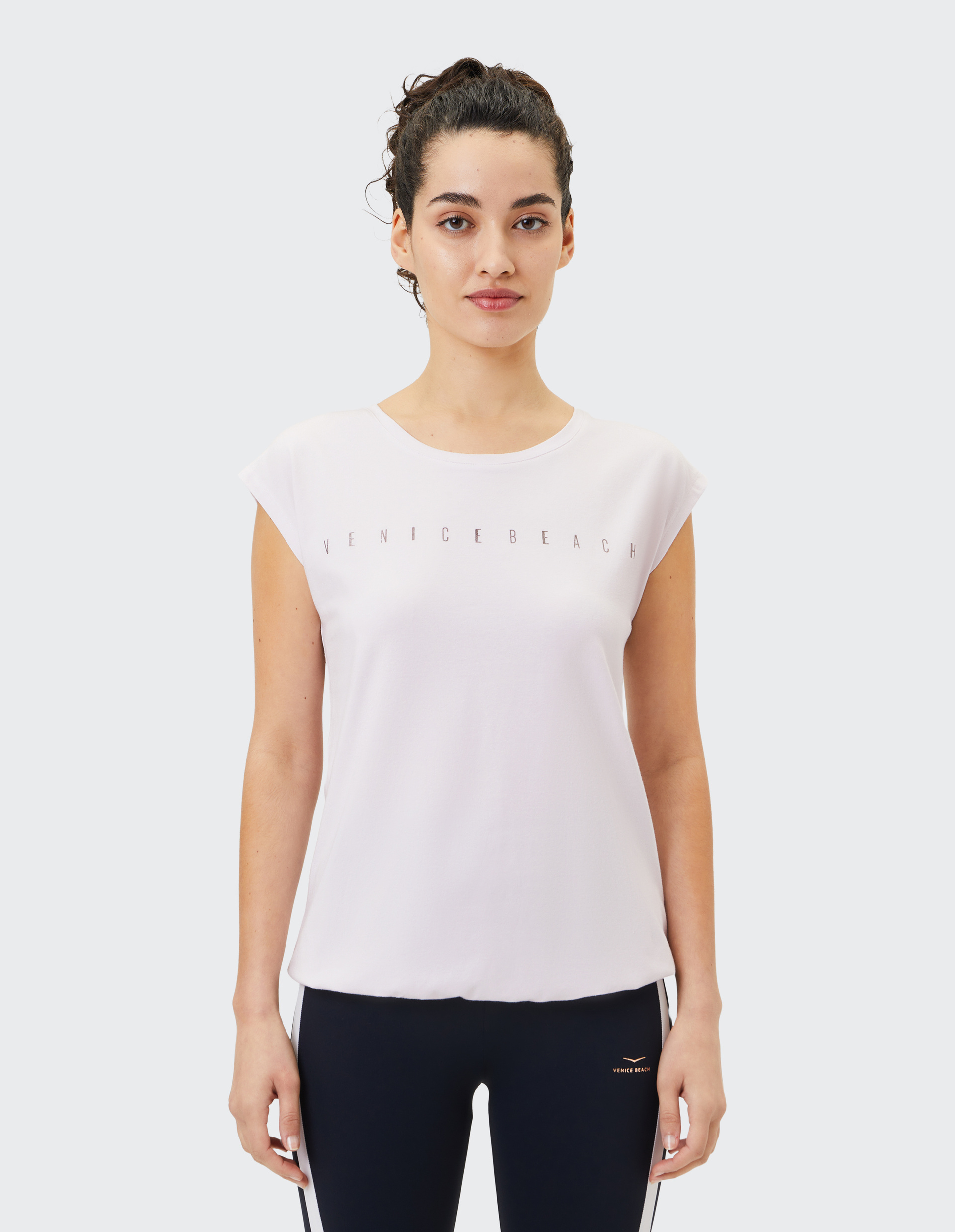 Frau mit lockigem Haar trägt weißes Venice Beach T-Shirt und dunkle Leggings, steht frontal vor hellem Hintergrund.