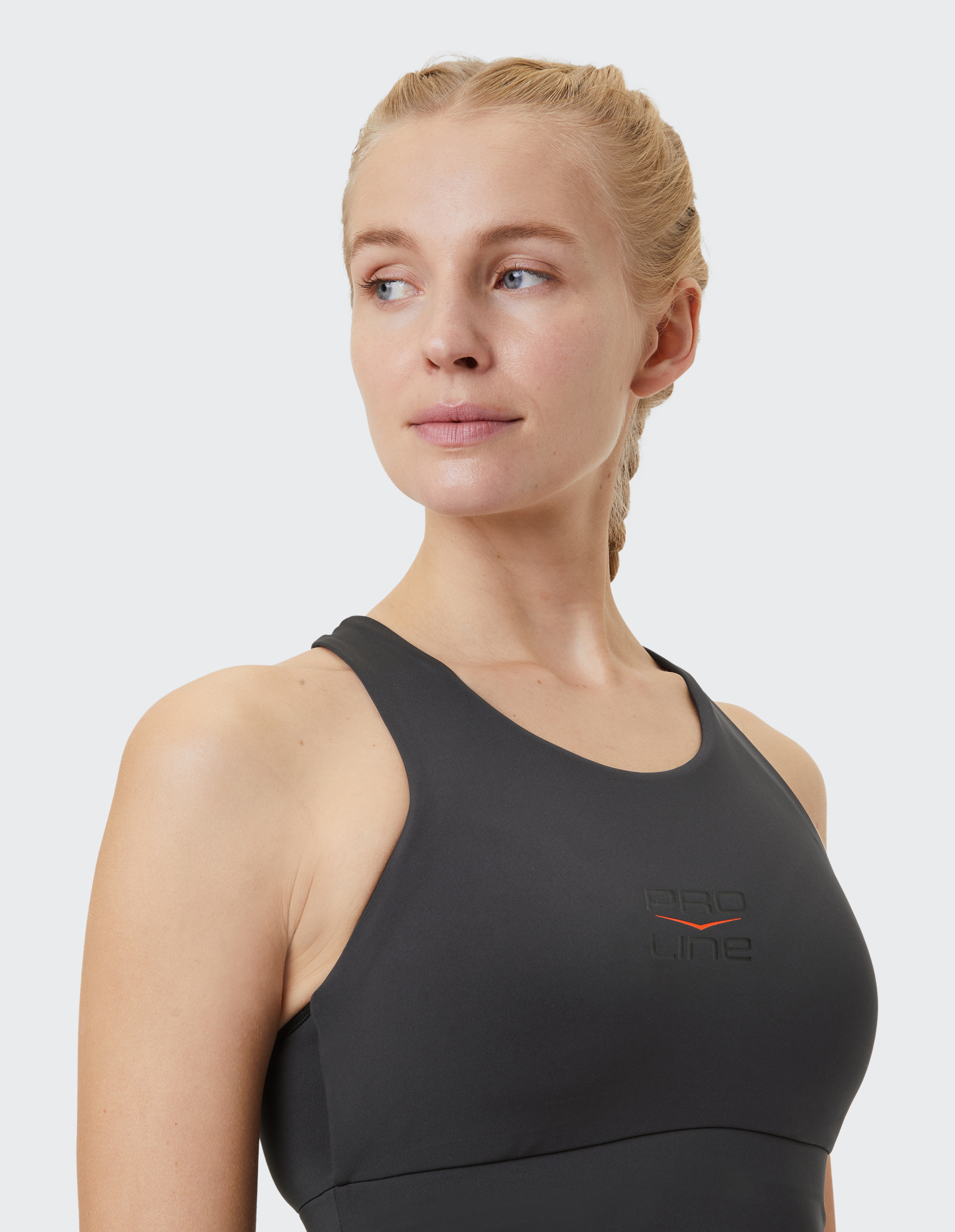 Frau mit geflochtenem Haar in einem schwarzen Sporttop, schaut nach links, vor schlichtem, hellem Hintergrund.
