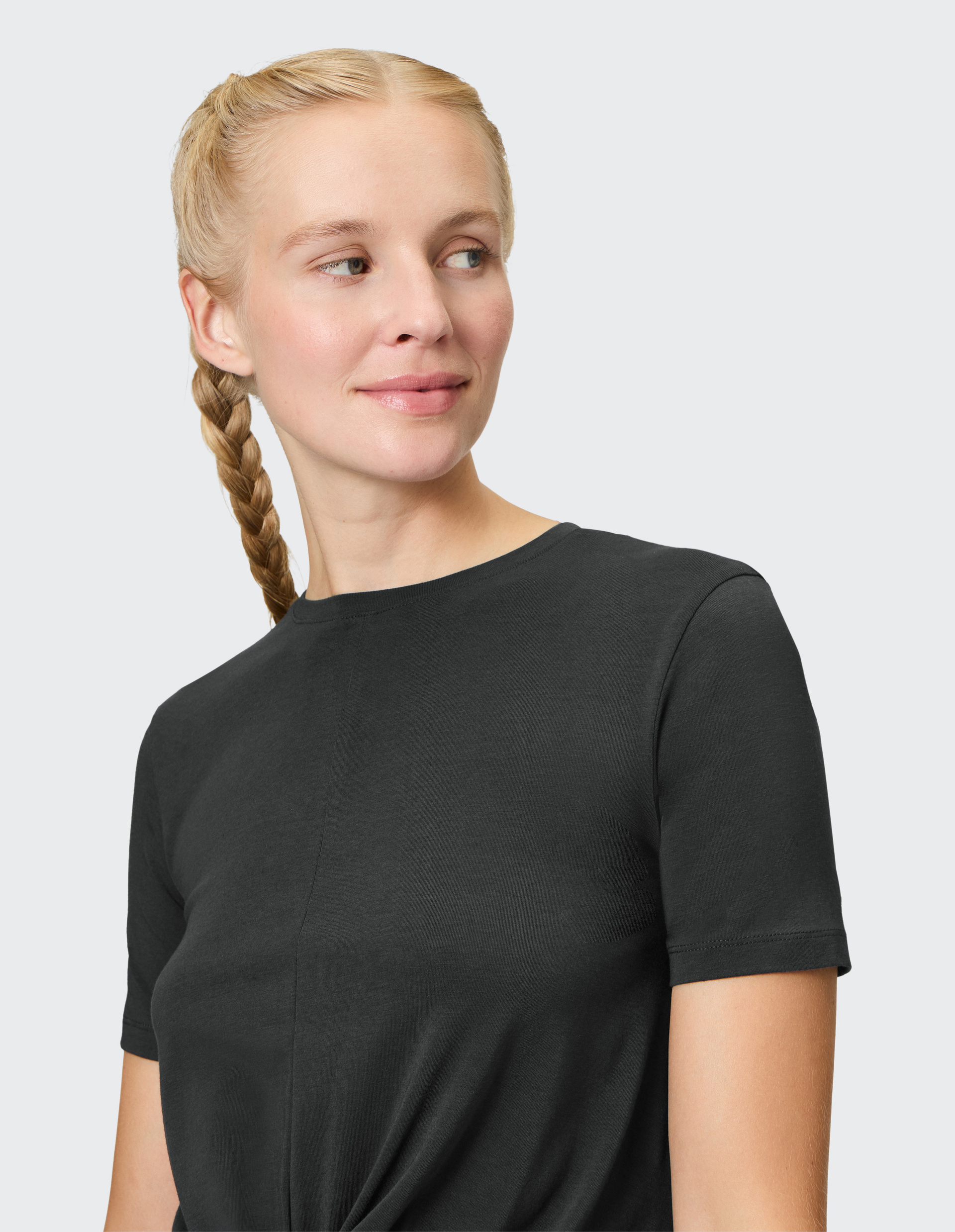 Eine lächelnde Person mit geflochtenem blonden Zopf trägt ein schwarzes T-Shirt und blickt nach links.