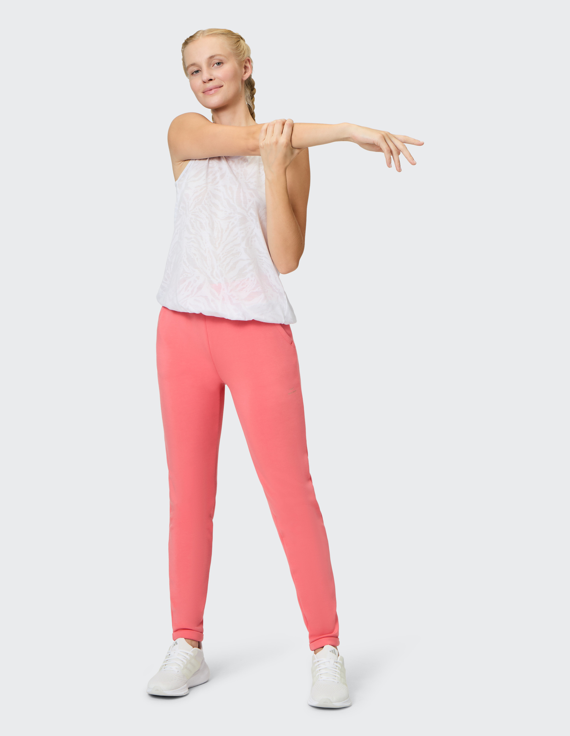 Eine Frau in rosa Hose und weißem Top dehnt ihren Arm und lächelt, die Haare sind geflochten; sie trägt weiße Schuhe.