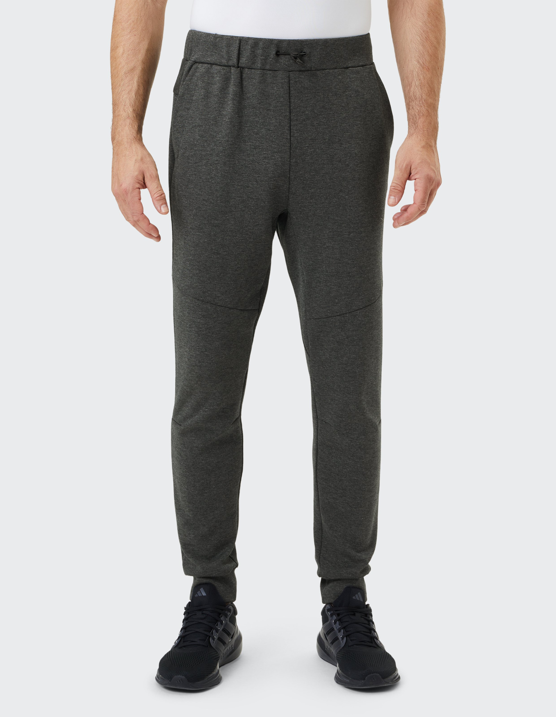 Mann in weißem Shirt trägt dunkelgraue Jogginghose und schwarze Turnschuhe, steht vor hellem Hintergrund, Hände locker an den Seiten.