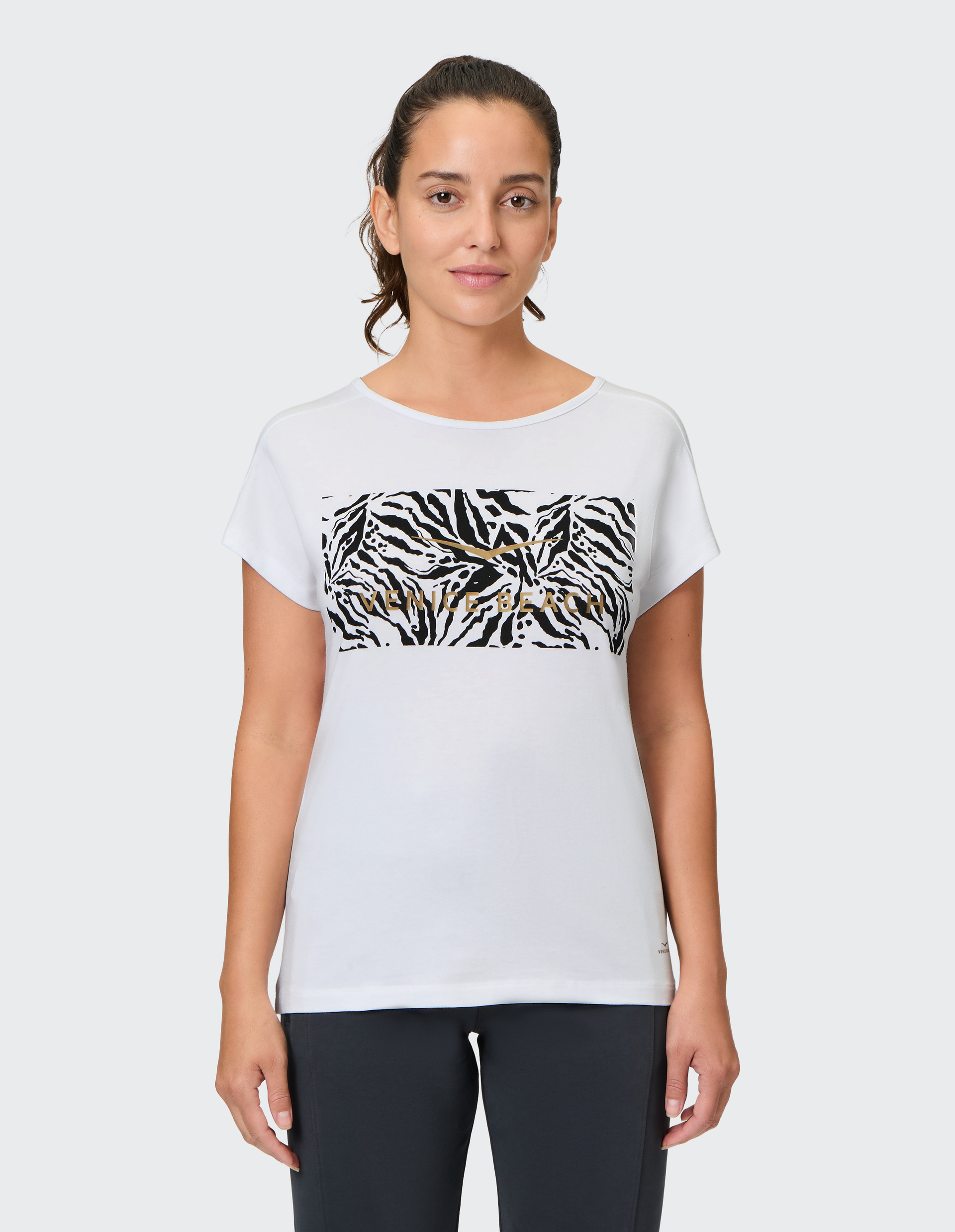 Frau in weißem T-Shirt mit Zebramuster-Print, kurzen Ärmeln; sie lächelt leicht, trägt dunkle Hose, Hintergrund einfarbig.