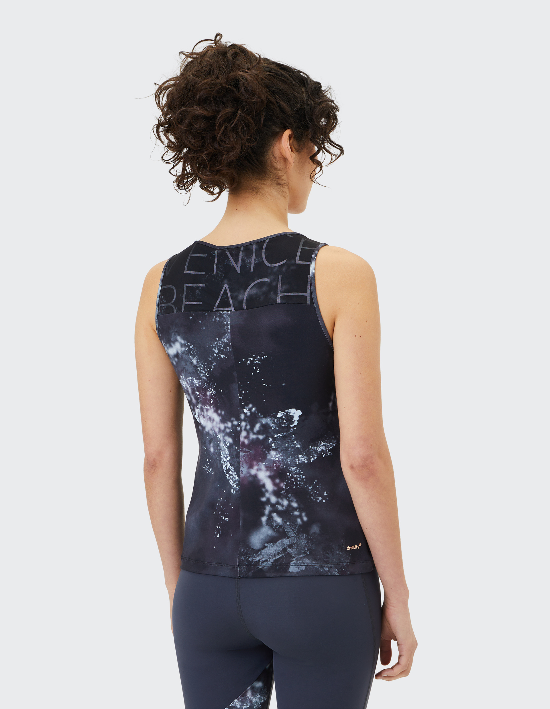 Frau mit lockigem Haar trägt ein ärmelloses Sporttop mit abstraktem Muster und VENICE BEACH-Schriftzug, passend zu dunklen Leggings.