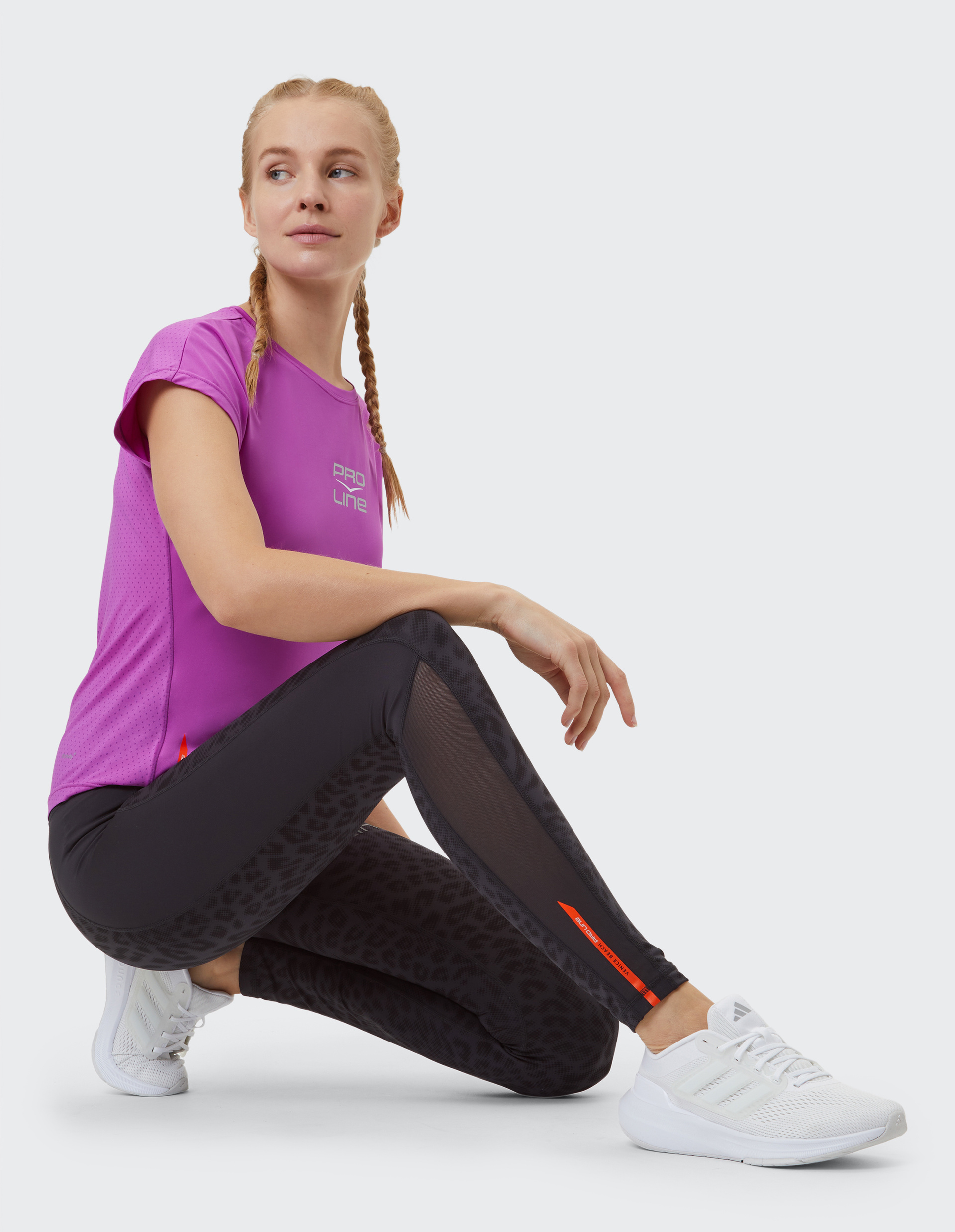 Frau in pinkem T-Shirt und schwarzer Leggings mit Muster kniet auf dem Boden, trägt weiße Sneaker und schaut zur Seite.