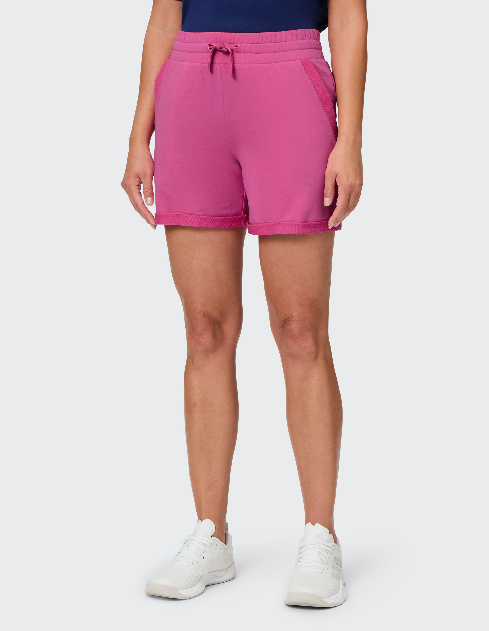 Person in pinker Shorts mit Kordelzug und dunkelblauem Oberteil, trägt weiße Turnschuhe, vor grauem Hintergrund stehend.