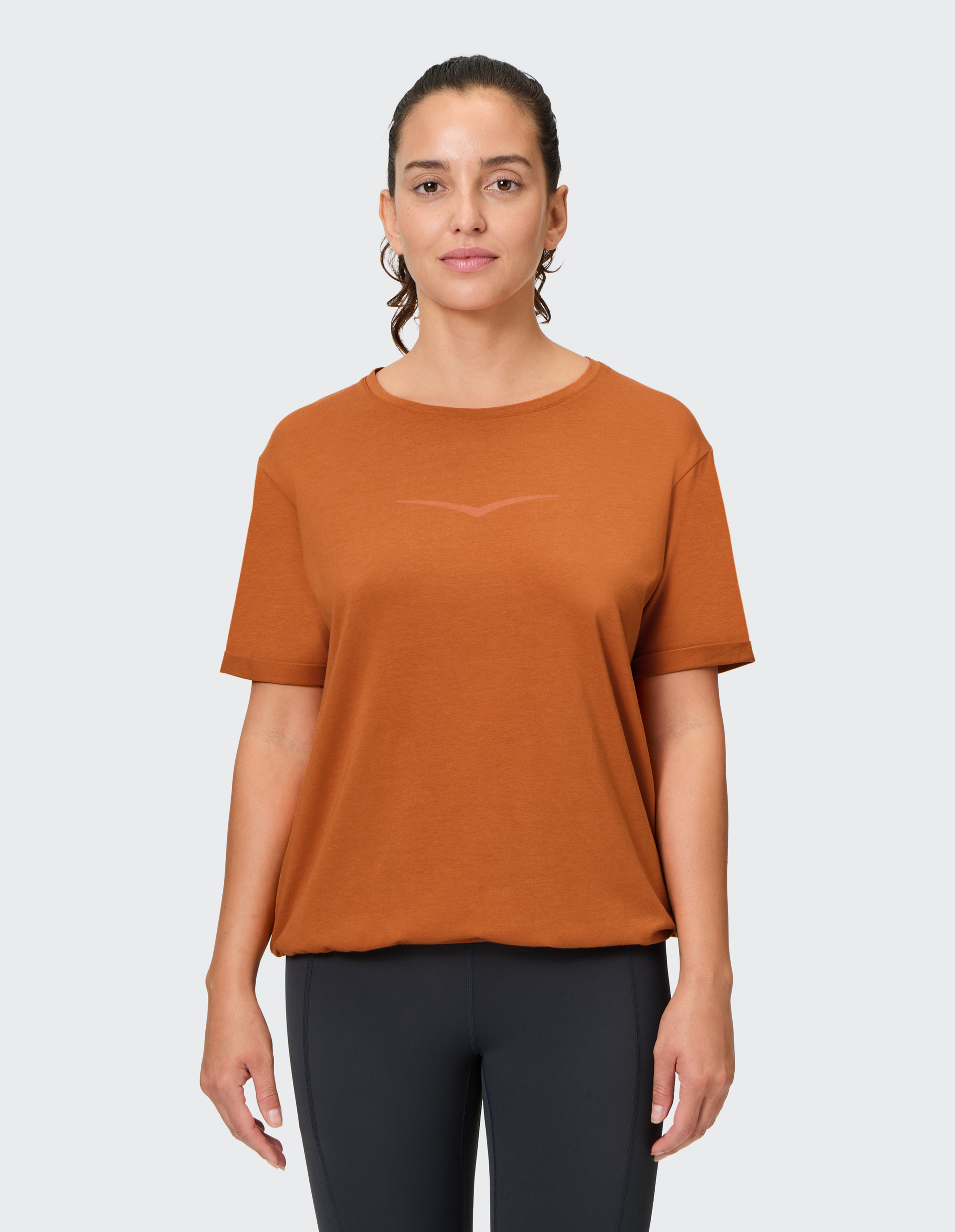 Eine Frau in einem orangefarbenen T-Shirt und schwarzen Leggings steht vor einem hellgrauen Hintergrund und schaut geradeaus.