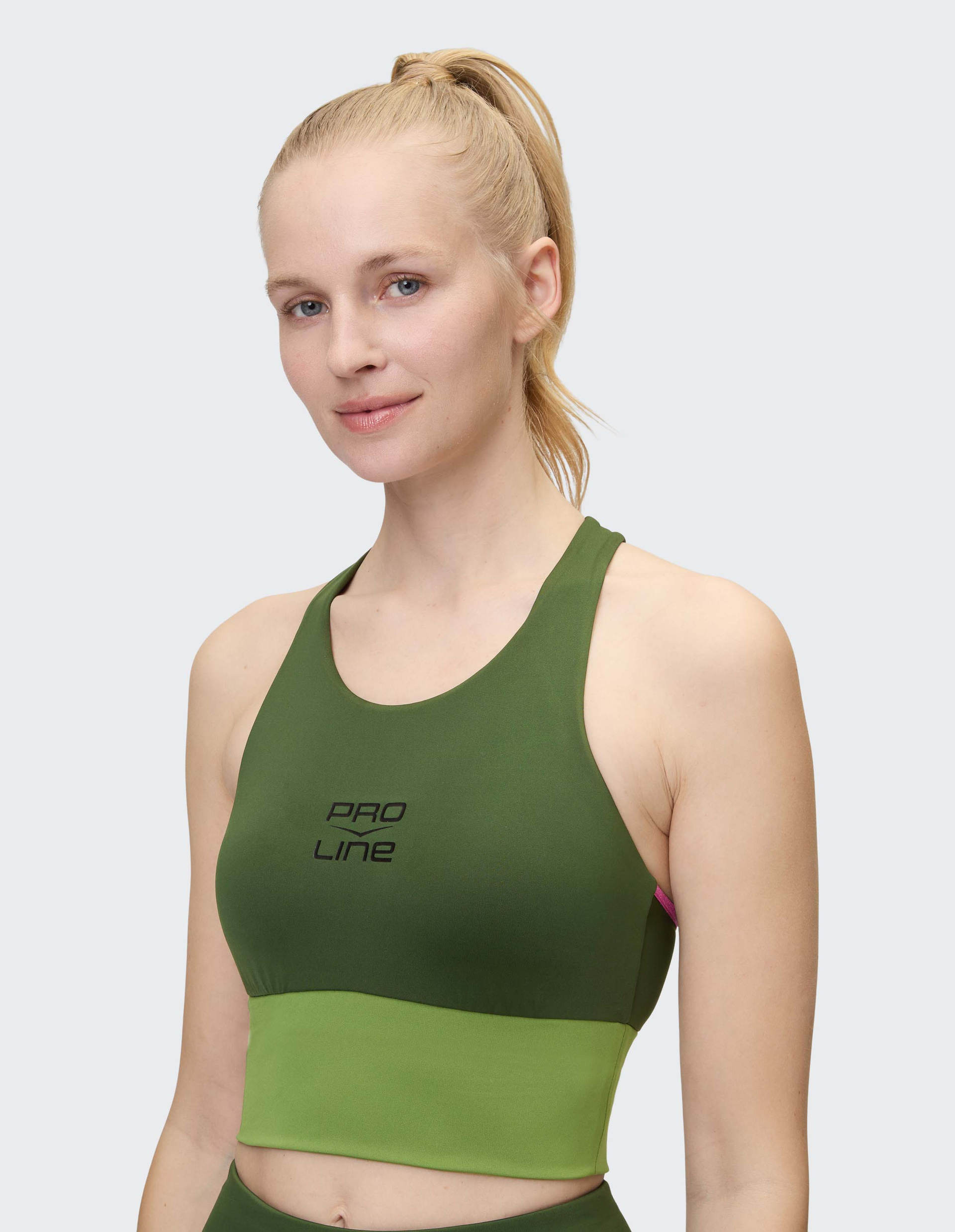 Eine Frau mit blonden Haaren im Pferdeschwanz trägt ein zweiteiliges, grünes Sportoutfit mit der Aufschrift "Pro Line".