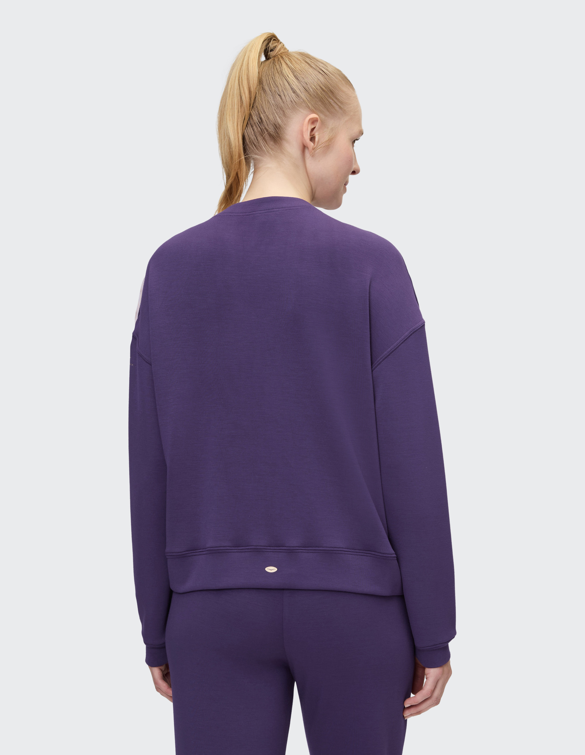 Eine Person mit blondem Pferdeschwanz trägt von hinten gesehen einen lilafarbenen Trainingsanzug mit langärmeligem Oberteil.