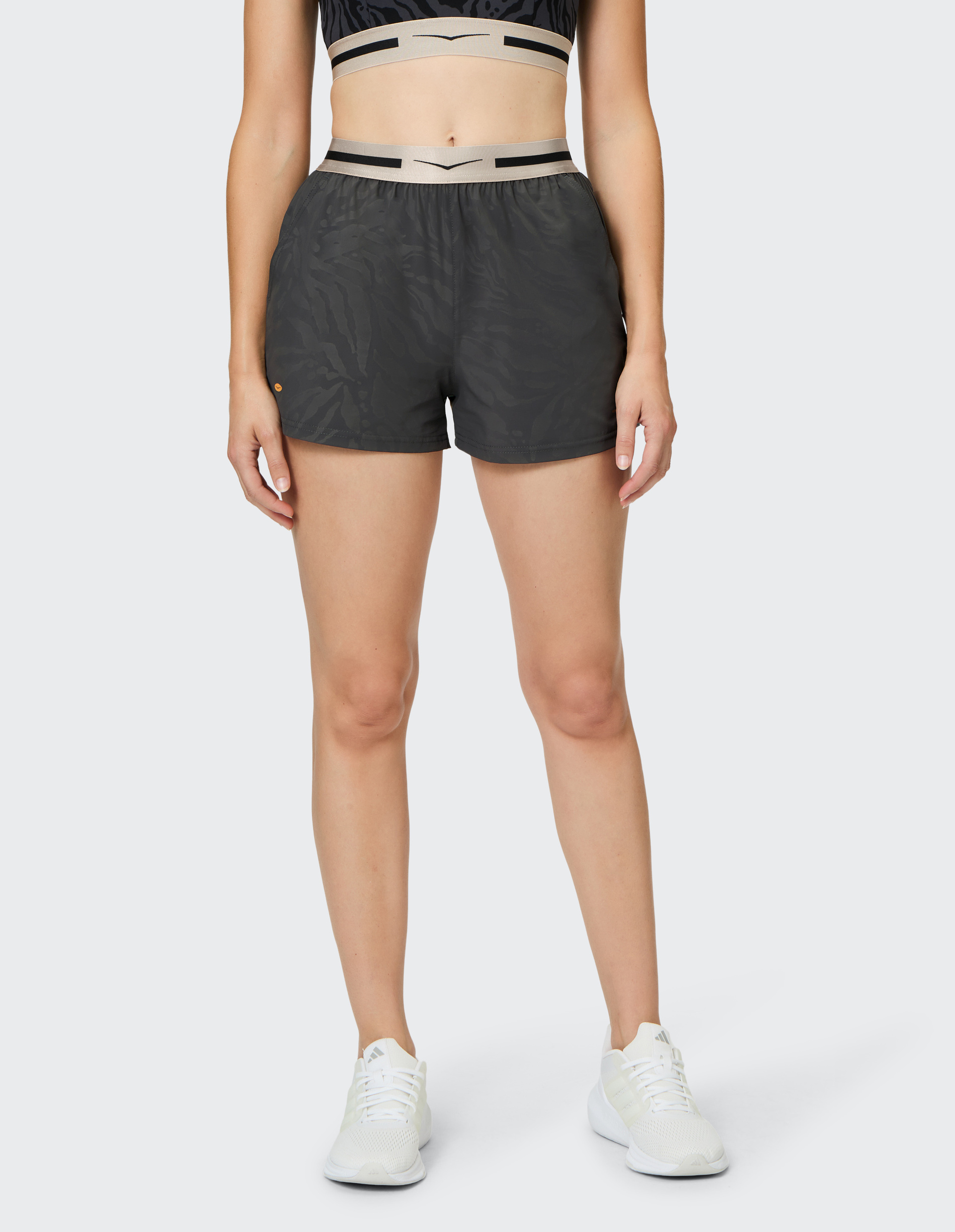 Eine Frau trägt schwarze Shorts mit camouflagemuster, ein passendes Top und weiße Sneakers, stehend vor einem neutralen Hintergrund.