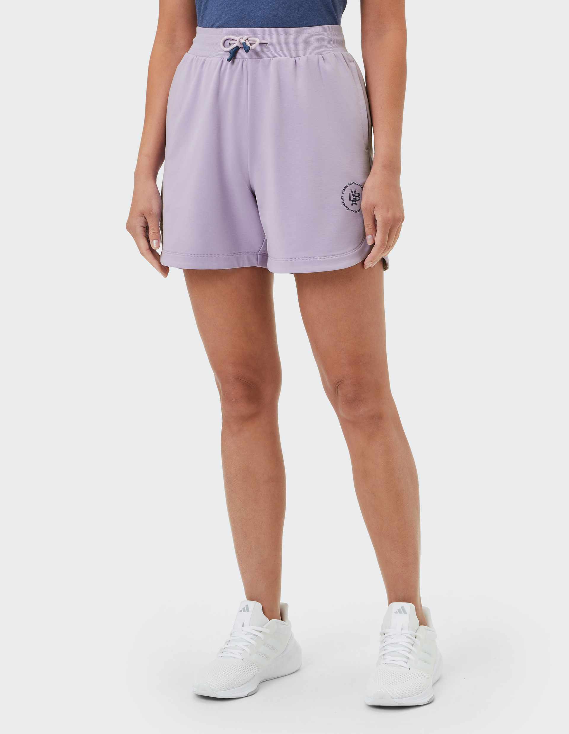 Frau trägt fliederfarbene Shorts mit Kordelzug und Logo, dazu weißes T-Shirt und Turnschuhe vor hellem Hintergrund.