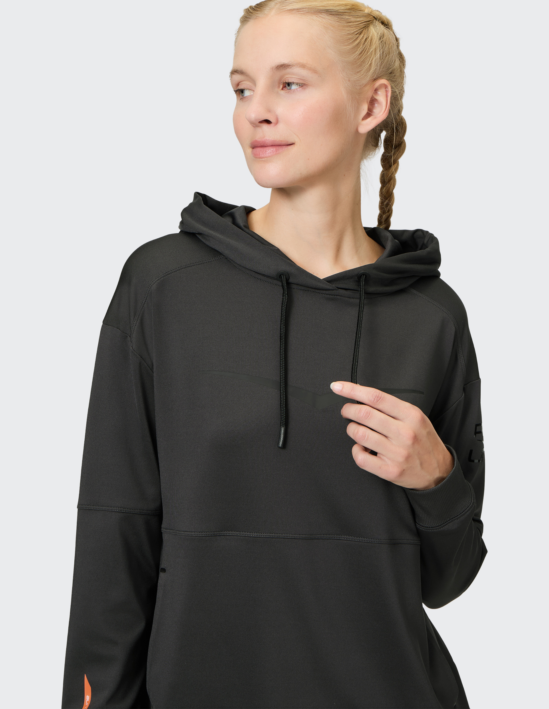 Frau mit geflochtenem Zopf trägt einen dunklen Hoodie und blickt lächelnd zur Seite; sie hält eine Kordel des Hoodies.