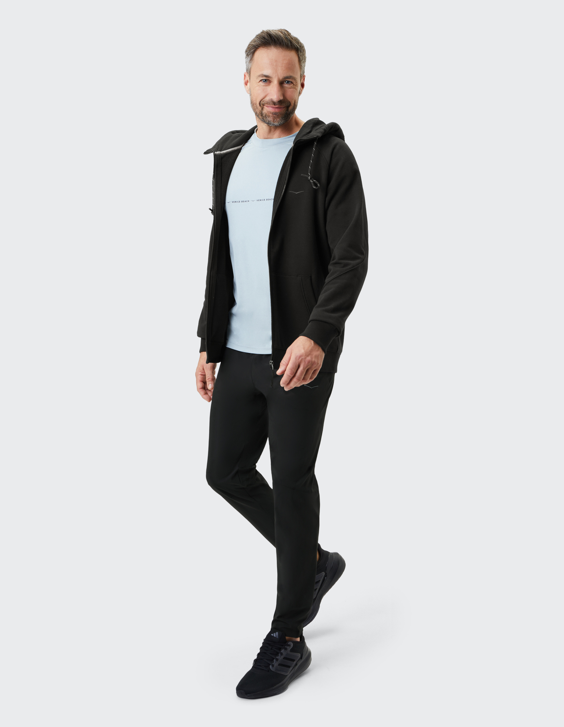 Mann mit Bart trägt schwarze Kapuzenjacke, hellblaues Shirt und schwarze Hose, lächelt und steht auf hellgrauem Hintergrund.