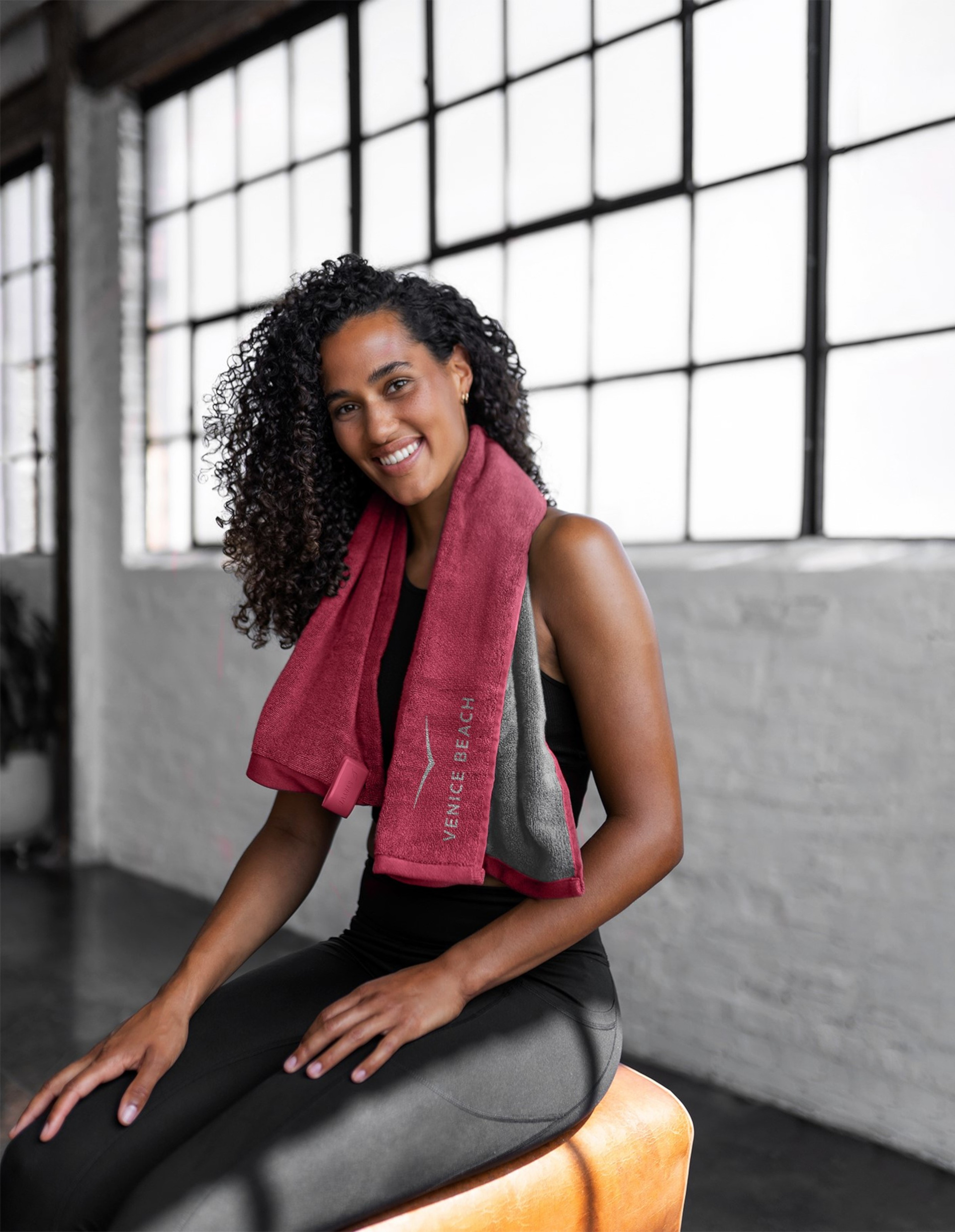 Frau mit lockigem Haar sitzt lächelnd in Sportkleidung auf einer Bank, ein rotes Handtuch um den Nacken gelegt, vor großen Fenstern.