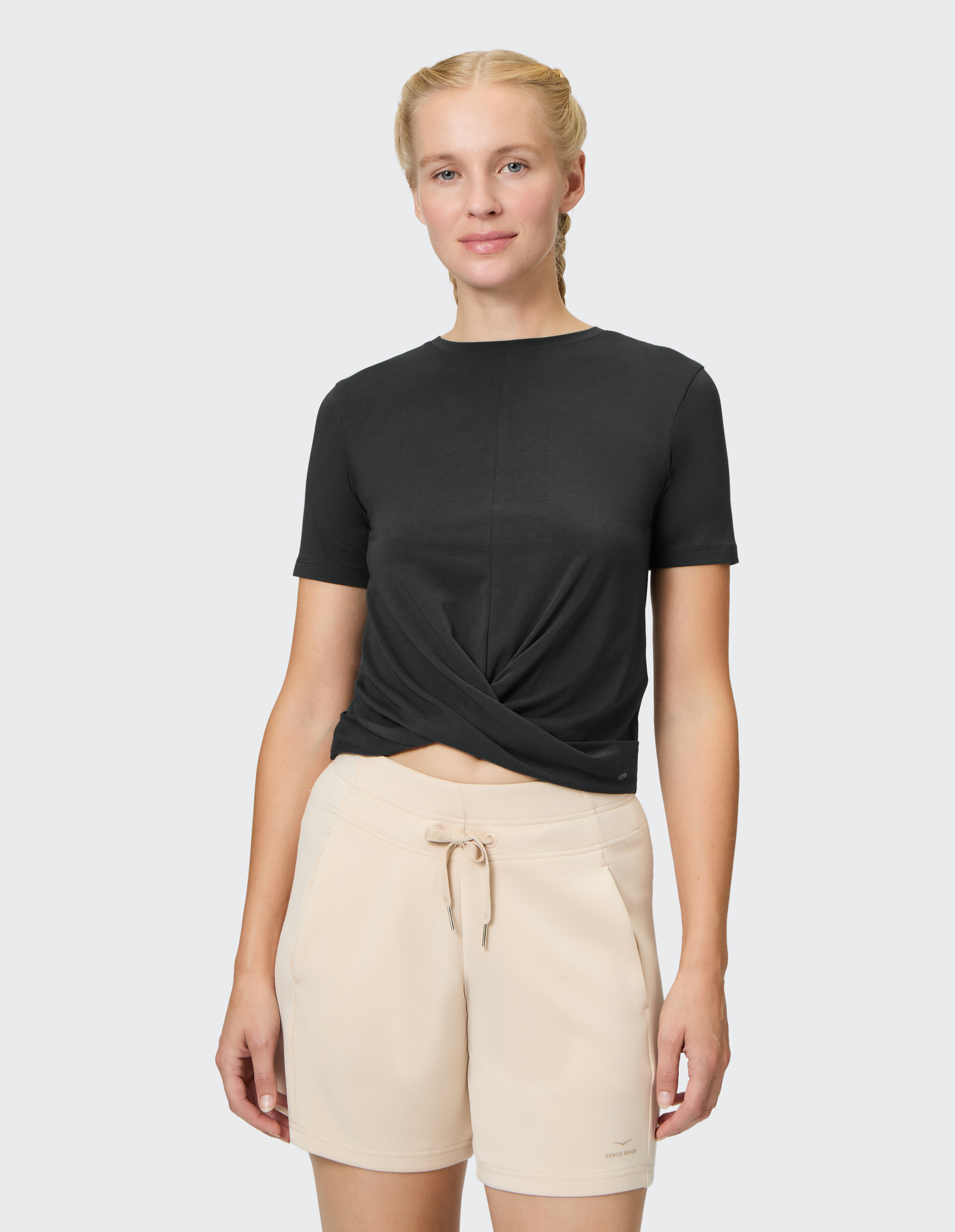 Frau trägt schwarzes T-Shirt und beige Shorts mit Kordelzug vor hellem Hintergrund, lächelt leicht mit geflochtenem Haar.