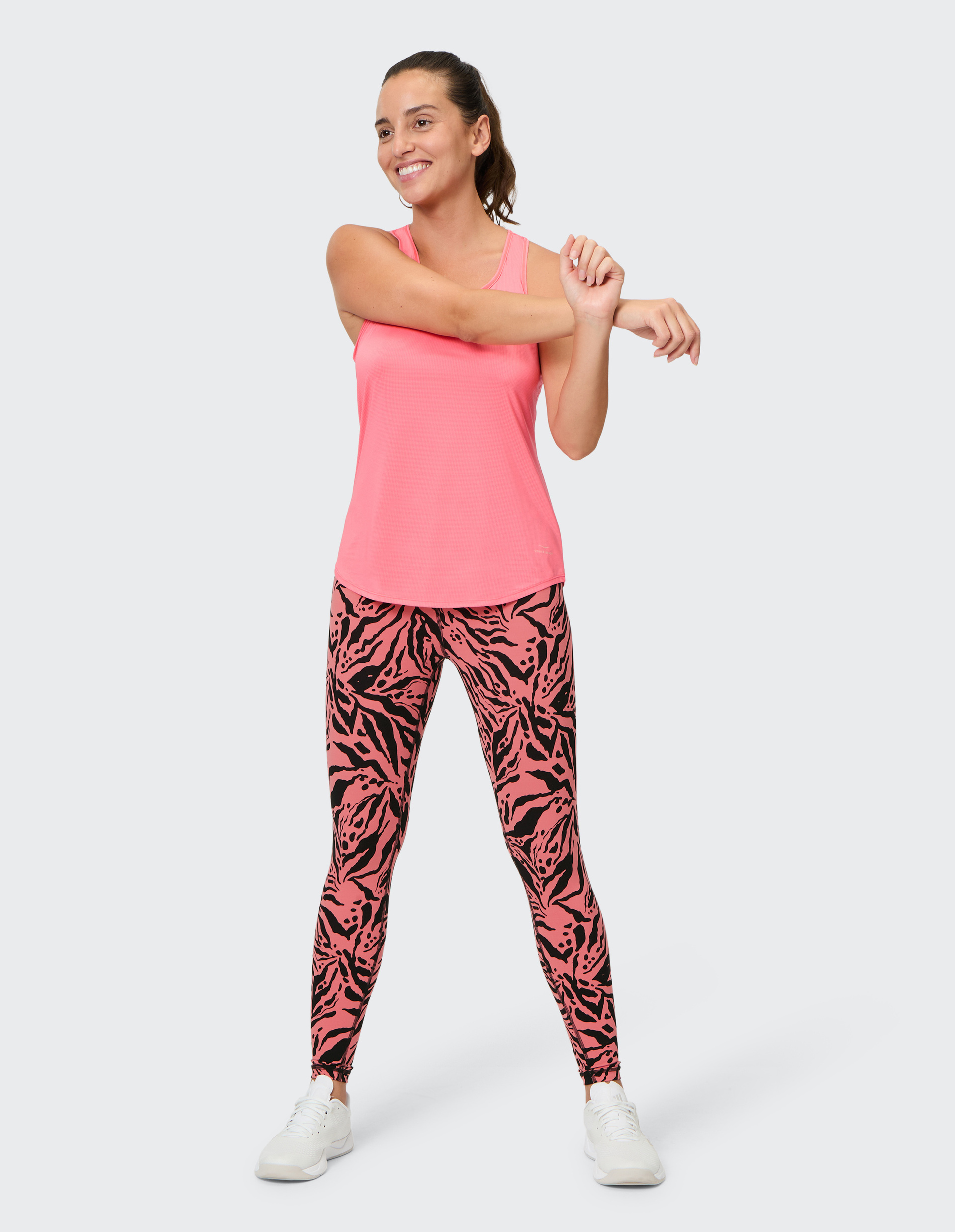 Frau in pinkem Tanktop und gemusterten Leggings dehnt lächelnd ihren Arm vor neutralem Hintergrund.